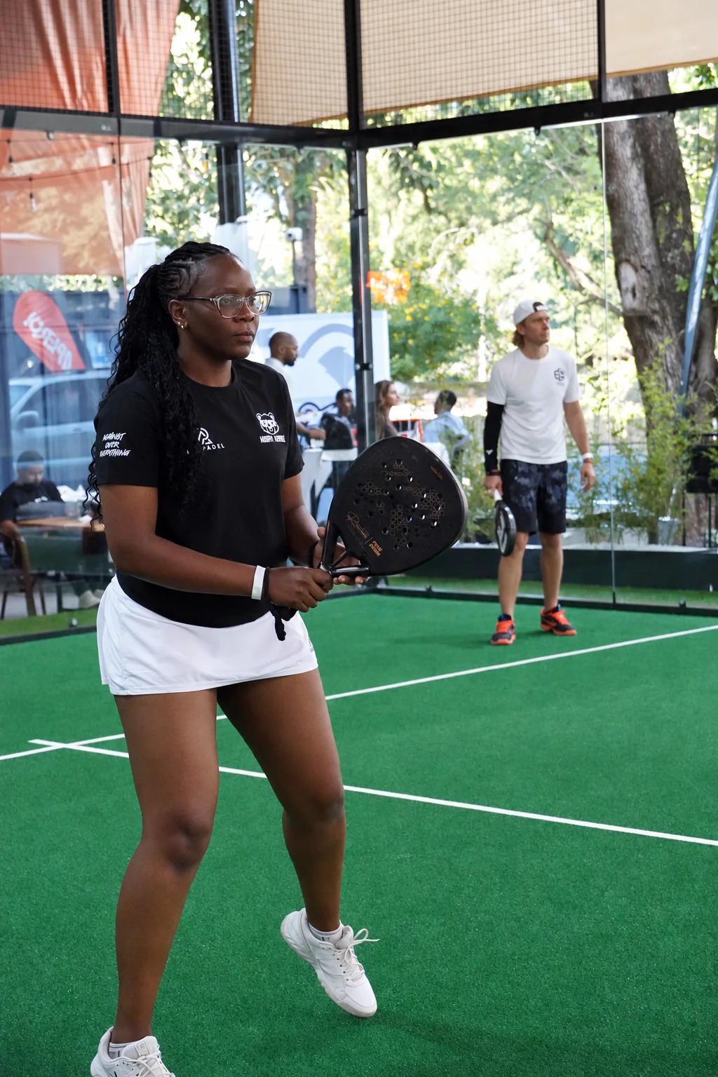Tonia on a green padel court in a black shirt and white skirt, holding her racket.